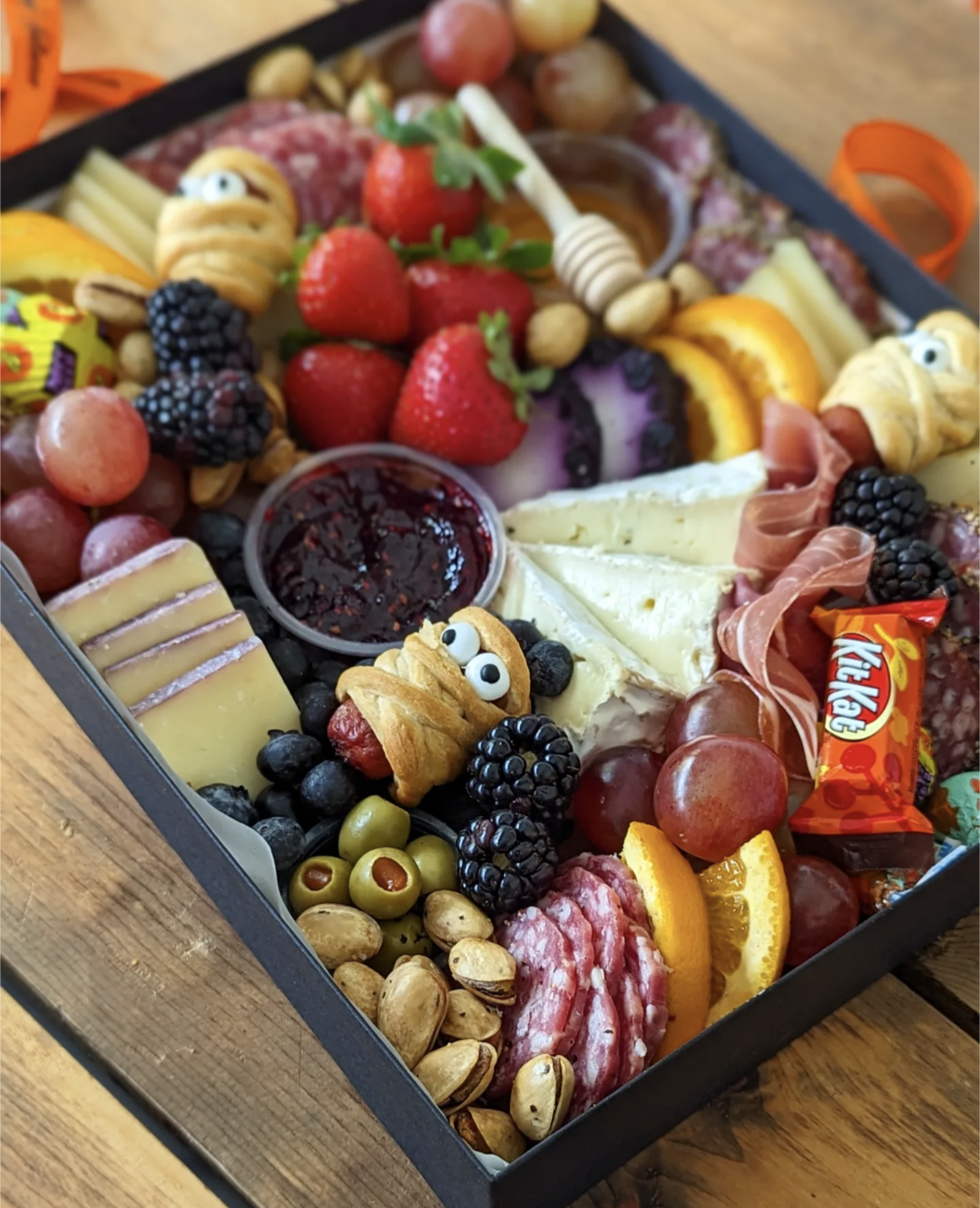 A platter of assorted Halloween-themed snacks including fake eyeballs, mummy sausages, molded cheeses, and various fruits and chocolates presented in a black box with 'Happy Halloween' ribbon.